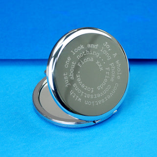 Silver compact mirror with personalised spiral message in typewriter font engraved on the front, shown in close up shot on blue background.